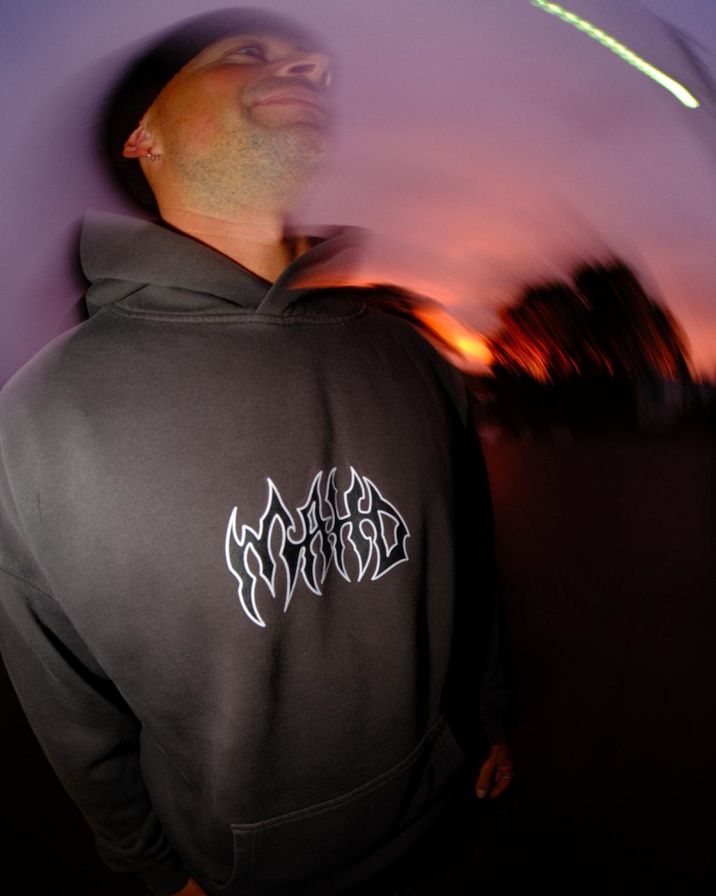 A person wears the Mahd Metal Hoodie—a gray hoodie with a stylized white logo—while standing outside at sunset, surrounded by a blurred, swirling visual effect.