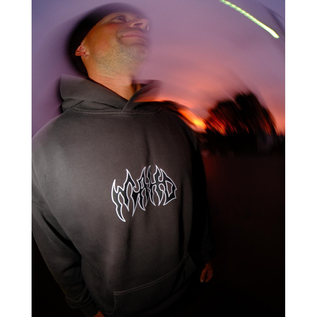 A person wears the Mahd Metal Hoodie—a gray hoodie with a stylized white logo—while standing outside at sunset, surrounded by a blurred, swirling visual effect.