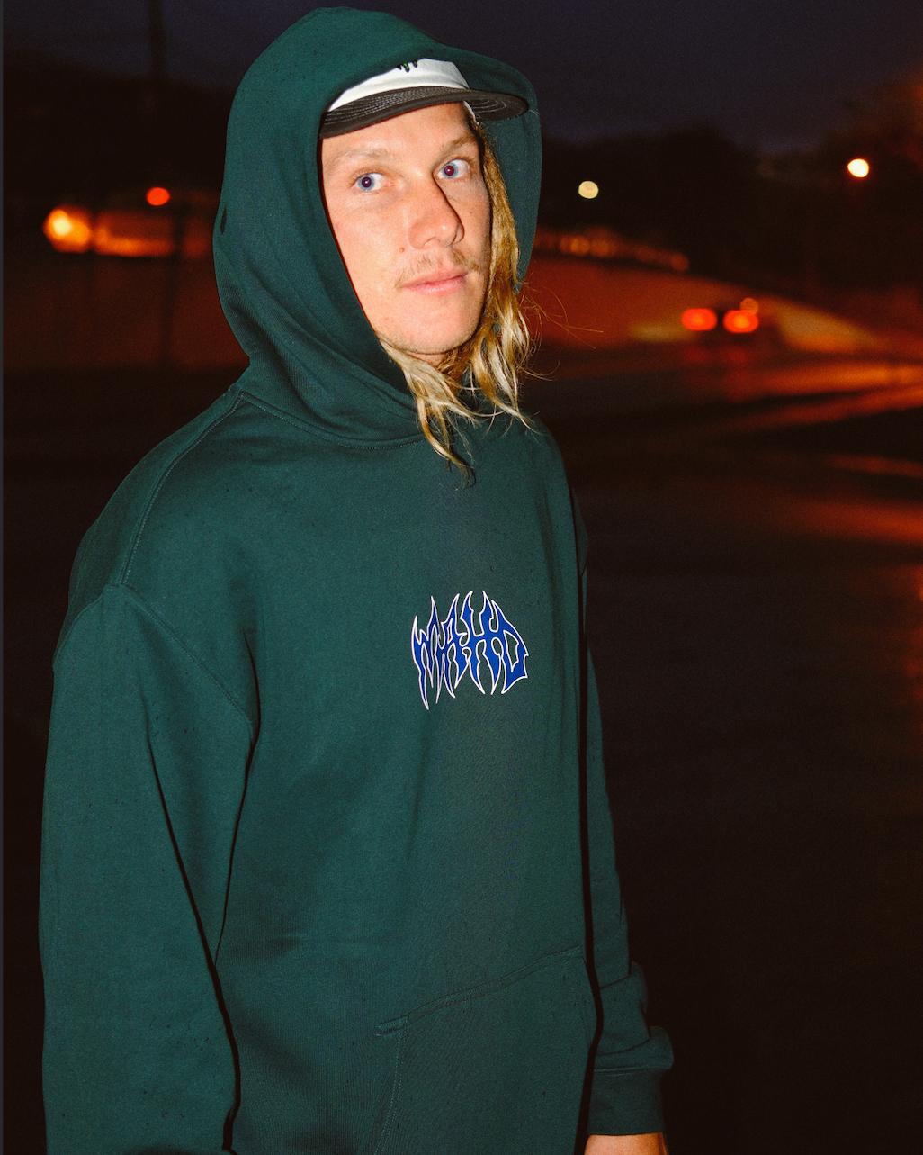 A person with long blond hair wears the Mahd Metal Hoodie and a cap, standing outside at night with blurred street lights in the background.