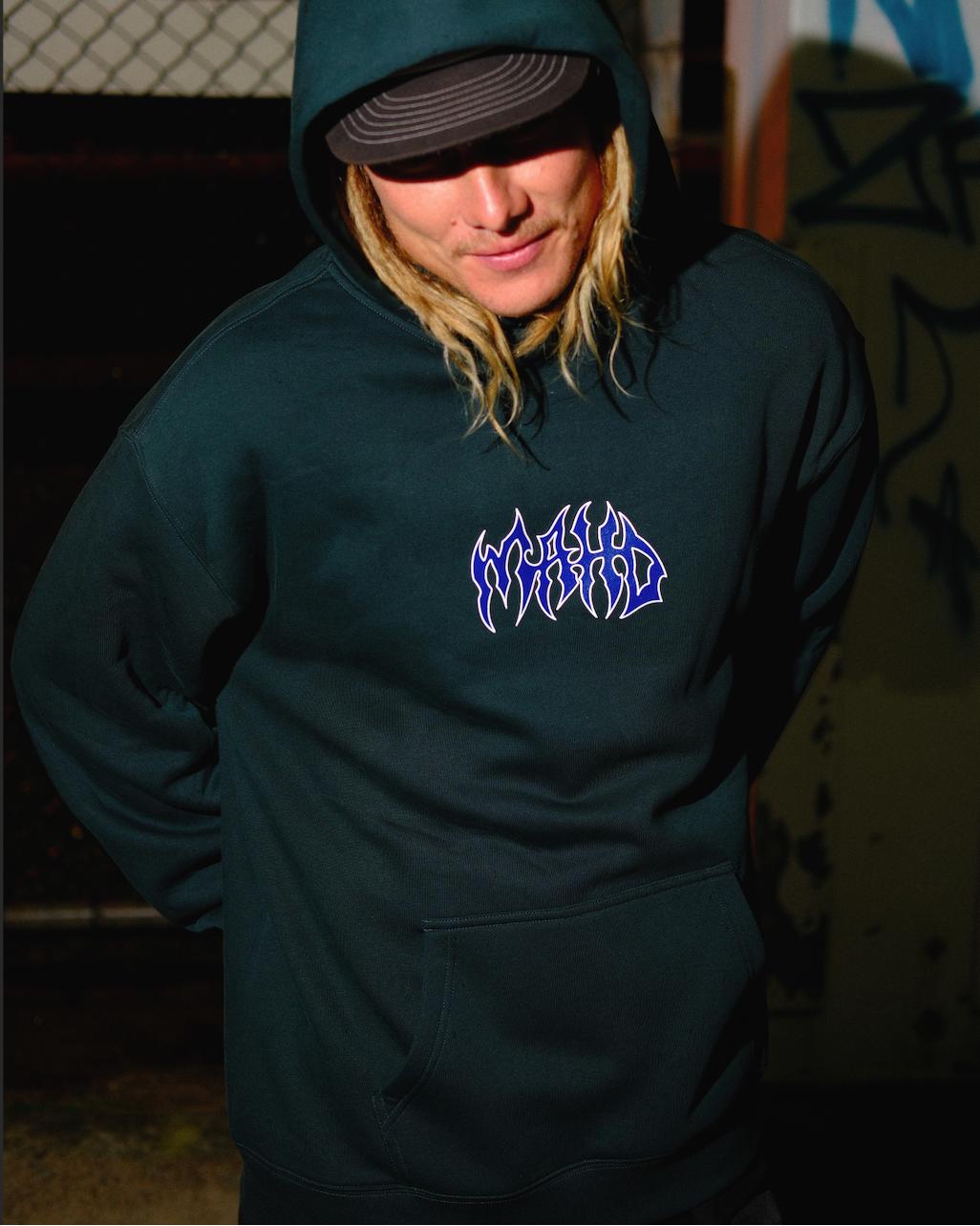 A person wearing the Mahd Metal Hoodie, featuring stylized "WORLD" text on the chest, stands outdoors at night by a chain-link fence with graffiti in the background.
