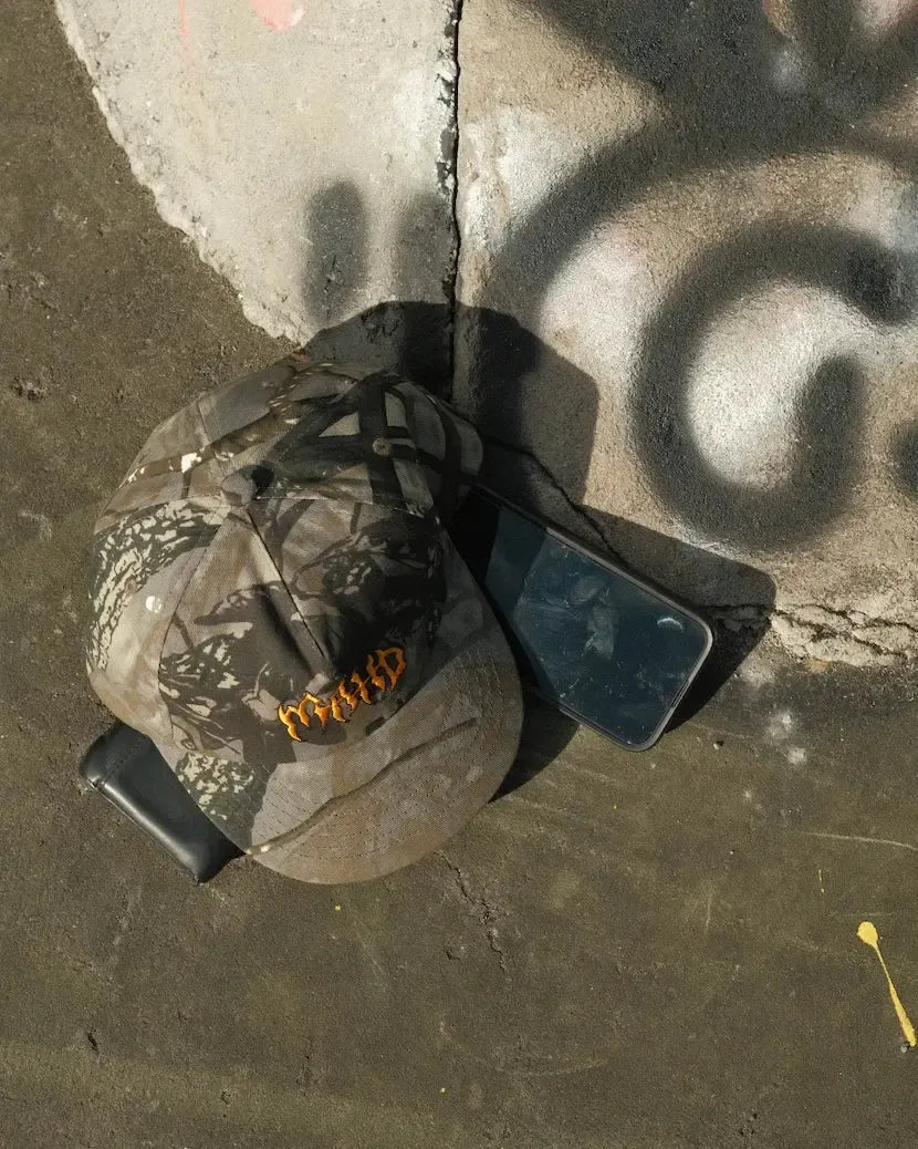 A Mahd - Metal Hat in a camouflage pattern rests on the ground next to two black smartphones—one with a cracked screen—by a sunlit concrete surface covered in graffiti.