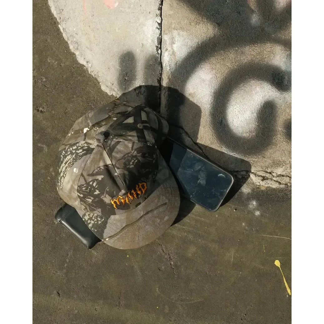 A Mahd - Metal Hat in a camouflage pattern rests on the ground next to two black smartphones—one with a cracked screen—by a sunlit concrete surface covered in graffiti.
