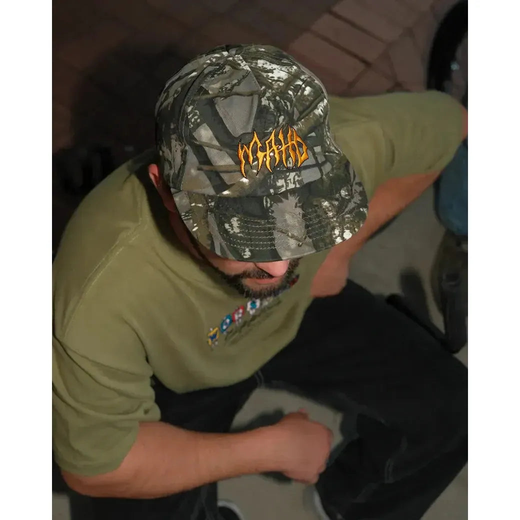 A person wearing the Mahd - Metal Hat, a green camouflage 5-panel cap with orange lettering, and an olive green shirt sits with their head slightly tilted down.