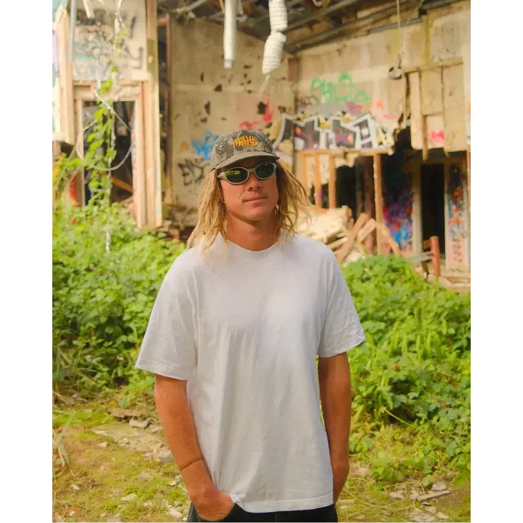 A person in a white t-shirt, sunglasses, and the Mahd - Metal Hat stands outdoors in front of an abandoned, graffiti-covered building surrounded by overgrown greenery.