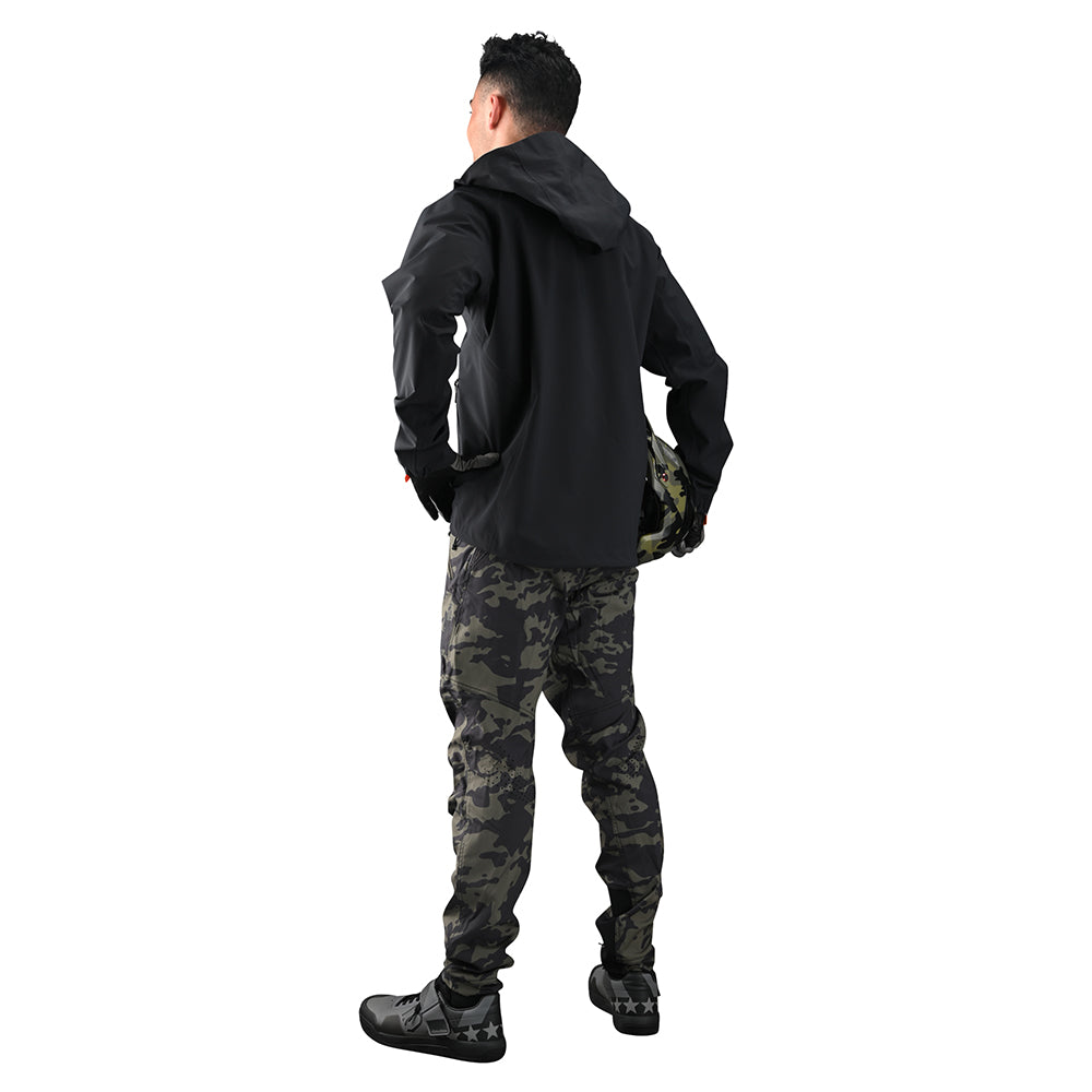 A person stands confidently with hands on hips, wearing the TLD Descent Jacket, a black waterproof hooded jacket, and camouflage pants.