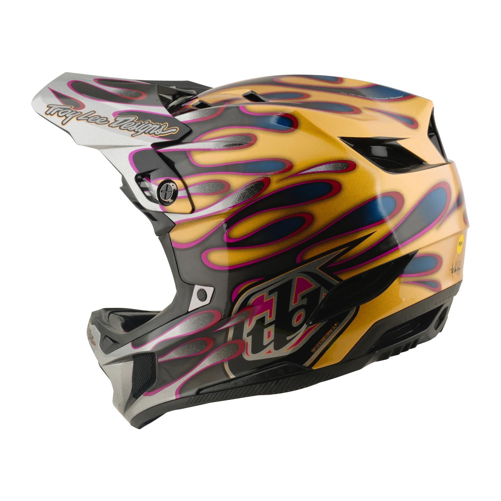 The TLD 26.1 D4 AS Carbon Helmet in Overspray Gold/Black features pink and blue flame graphics, a Troy Lee Designs visor logo, full-face coverage, advanced Mips protection, and is shown in side profile.