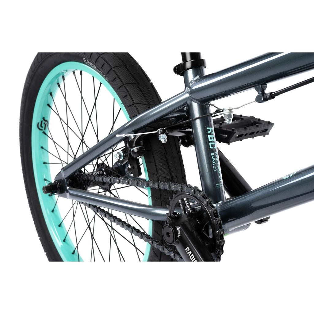 Close-up of the Radio Saiko 20 Inch Bike’s rear wheel, chain, pedals, and frame, highlighting Salt Tracer Tyres with black tread and teal rim on this metallic gray kids BMX bike.