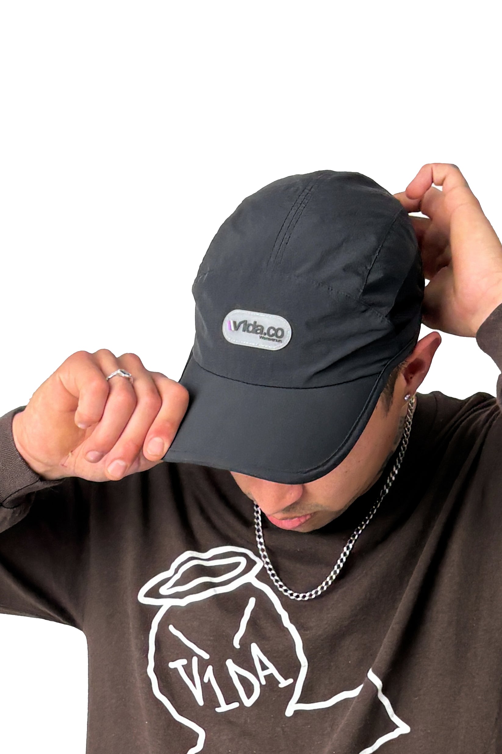 A person wearing the V1da We Never Run Cap with a "vida.co" logo and transpirable panel, a silver chain, and a brown shirt with a white graphic and "VIDA" text adjusts their cap against a white background.