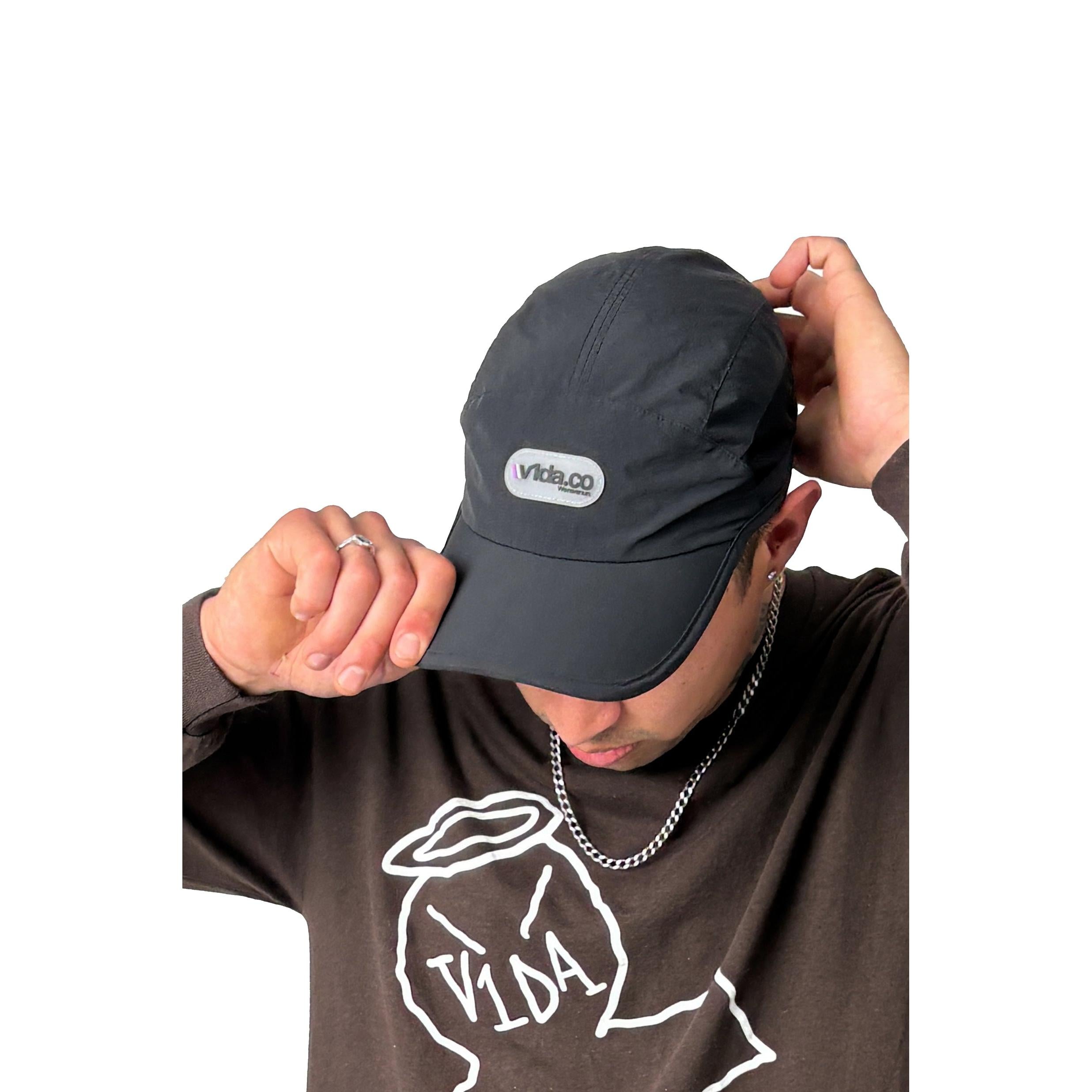 A person wearing the V1da We Never Run Cap with a "vida.co" logo and transpirable panel, a silver chain, and a brown shirt with a white graphic and "VIDA" text adjusts their cap against a white background.