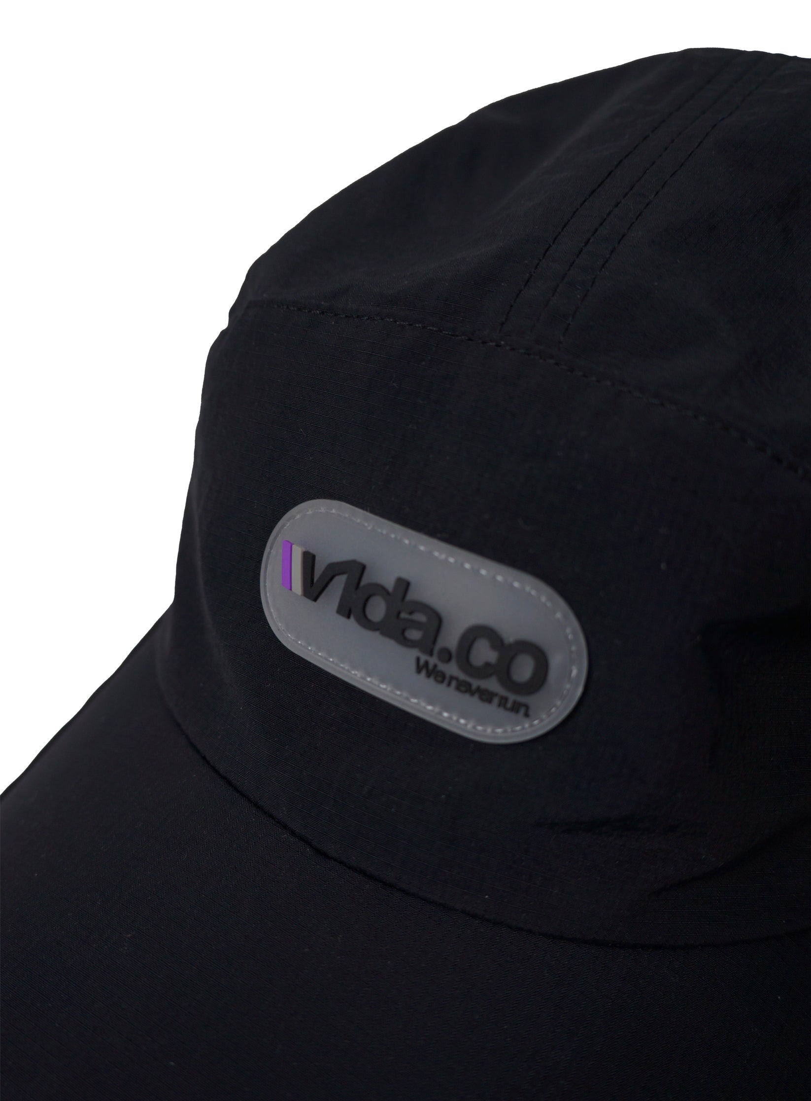 Close-up of the V1da We Never Run Cap, a black cap with a gray oval "Vida.CO Wanderism" patch, a small purple front stripe, and a breathable transpirable panel for comfort.