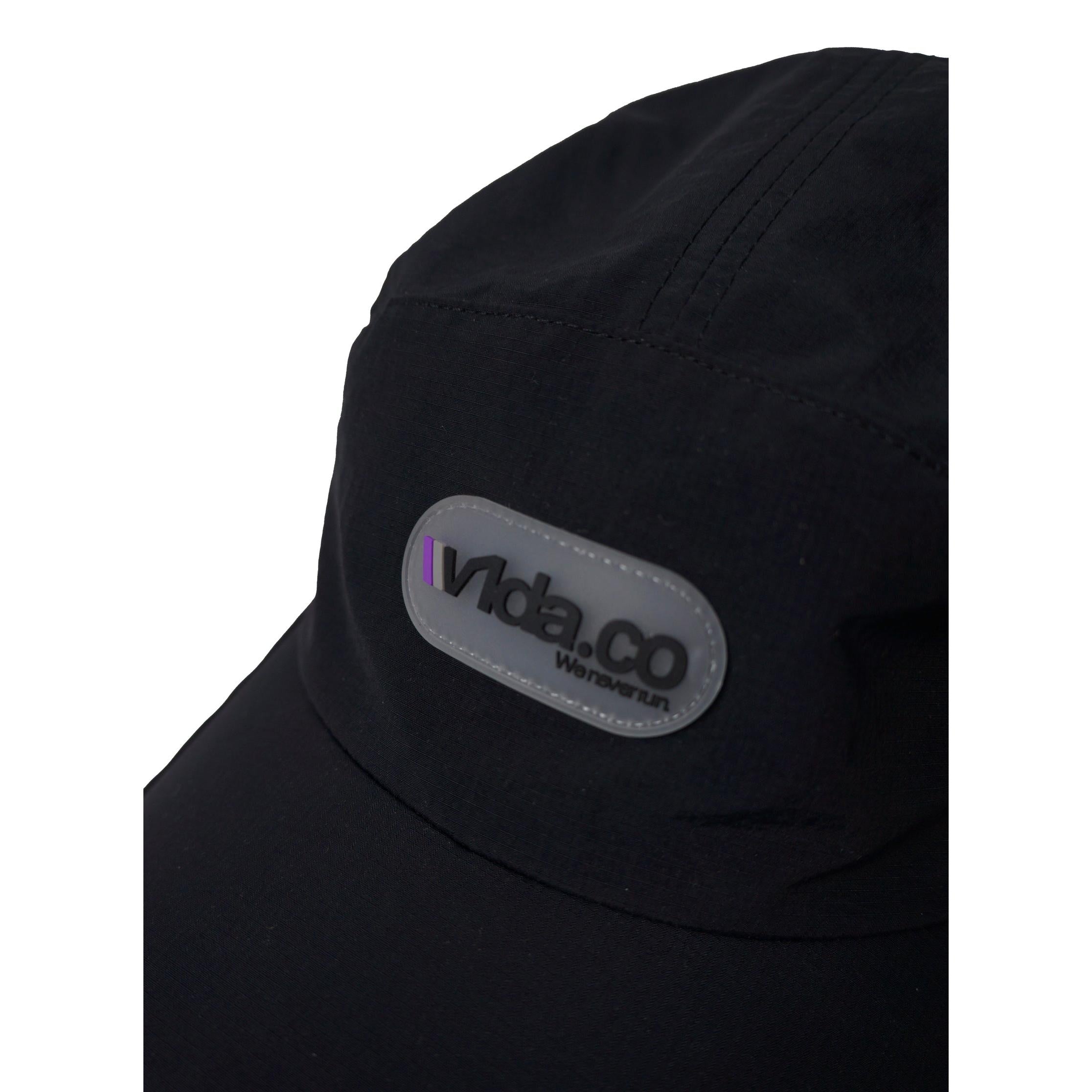 Close-up of the V1da We Never Run Cap, a black cap with a gray oval "Vida.CO Wanderism" patch, a small purple front stripe, and a breathable transpirable panel for comfort.