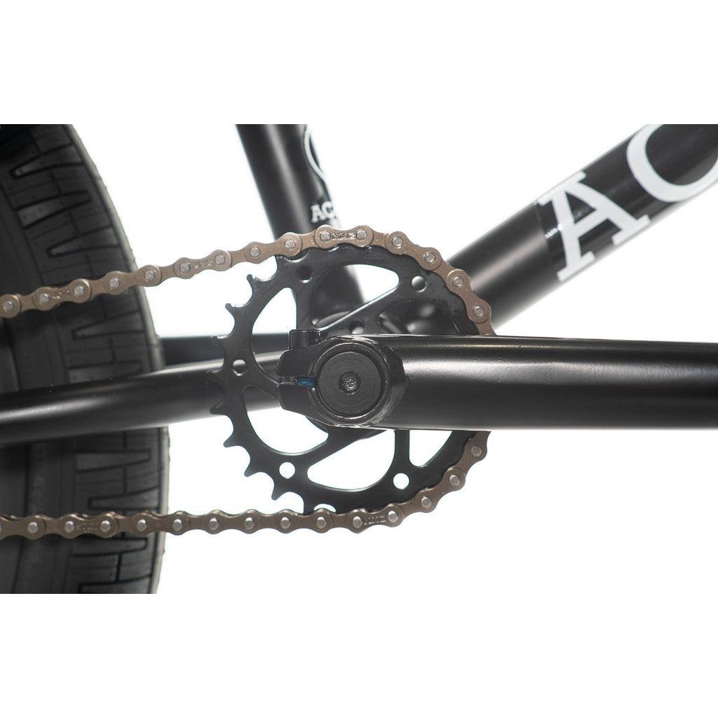 Close-up view of the freestyle BMX crankset on the Academy Entrant 20 Inch Bike, including the chain, chainring, and part of the tire and frame. The minimalist chainring design features evenly spaced holes, while the frame showcases a partial logo from Academy Bikes.