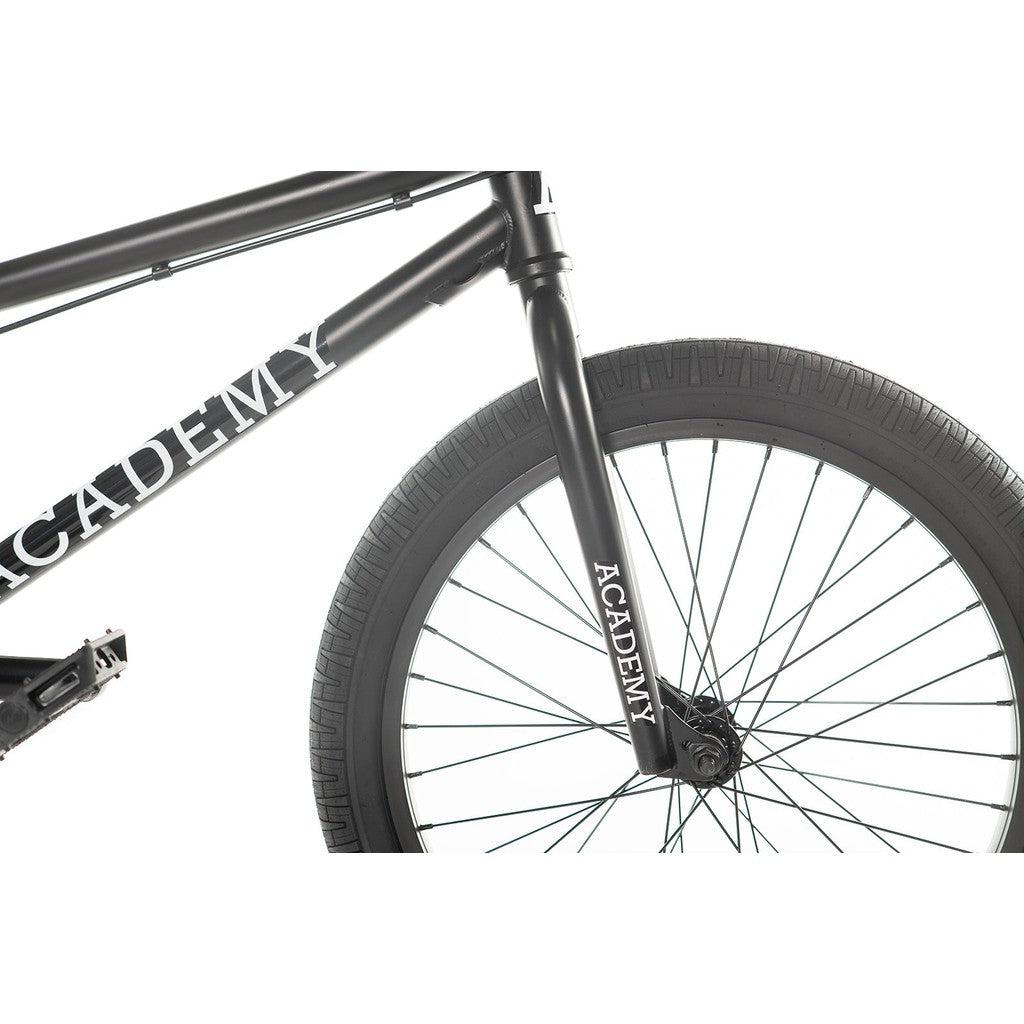 Close-up of the front tire and frame of an Academy Entrant 20 Inch Bike, showcasing the word "ACADEMY" printed on both the frame and fork. This entry-level BMX from Academy Bikes is matte black with shiny spokes, making it perfect for those new to freestyle BMX.