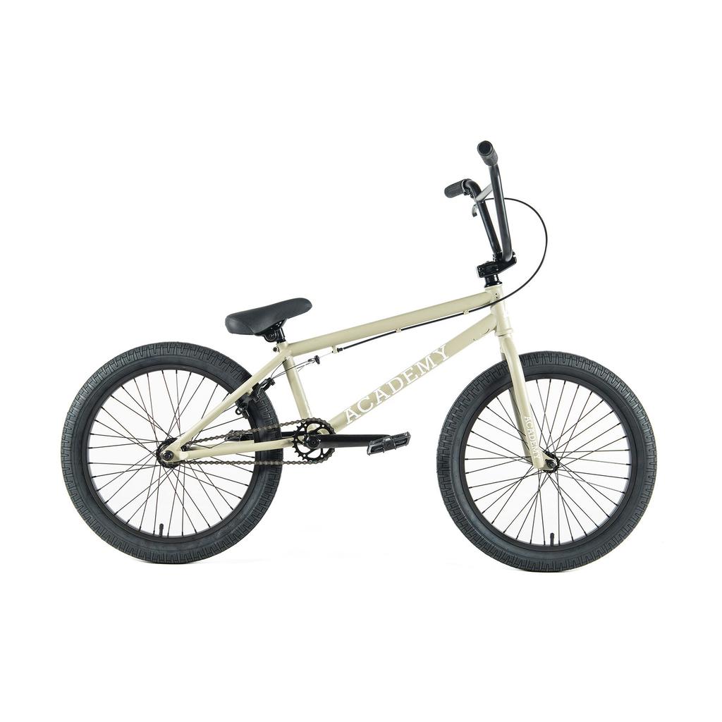 The Academy Entrant 20 Inch Bike is a beige entry-level freestyle BMX with black tires, black handlebars, and “ACADEMY” on the frame, shown against a white background.
