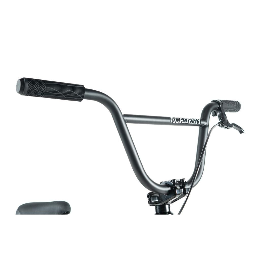 Close-up of Academy Inspire 18 Bike’s black handlebars with grips and brake lever, "Academy" branding on the crossbar, and double wall rims, all shown against a white background.