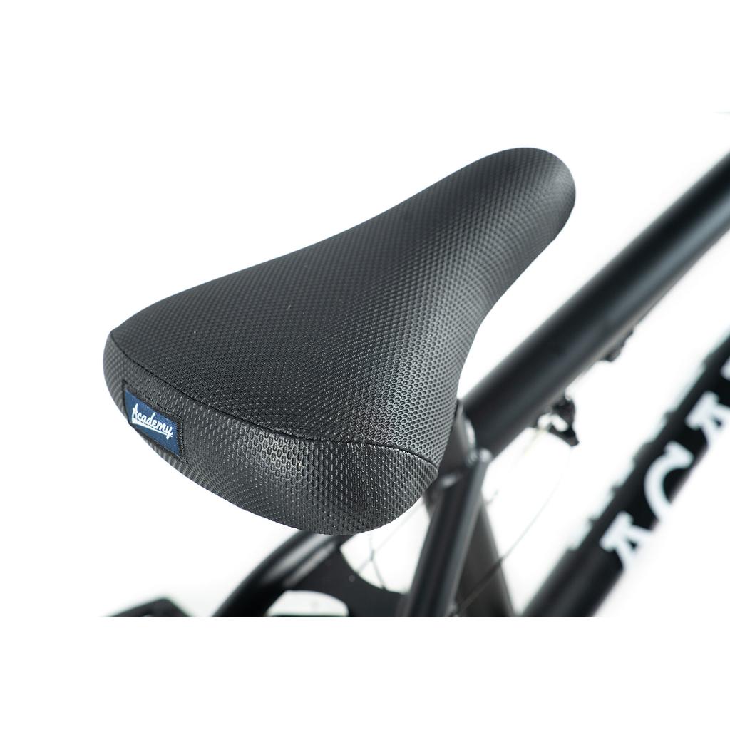 Close-up of a black textured BMX seat with a small blue "academy" label, mounted on the Academy Inspire 18 Bike’s black frame against a white background.