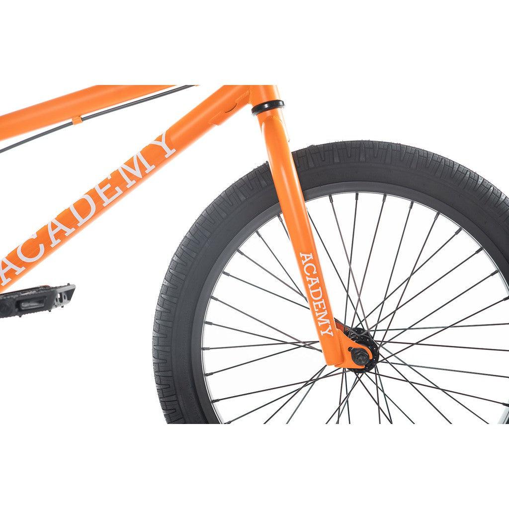 Close-up of the front section of an Academy Trooper 20 Inch Bike in orange, showcasing "ACADEMY" branding on its Hi-Ten steel frame and fork, accompanied by a black tire with sealed hubs and spokes.