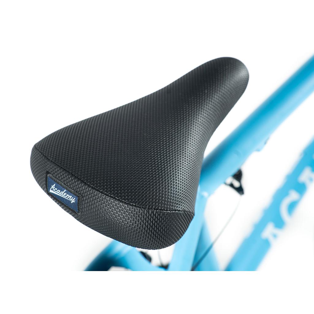 Close-up of a textured black seat with an "Academy" tag on the Academy Trooper 20 Inch Bike, shown attached to the entry-level blue BMX.
