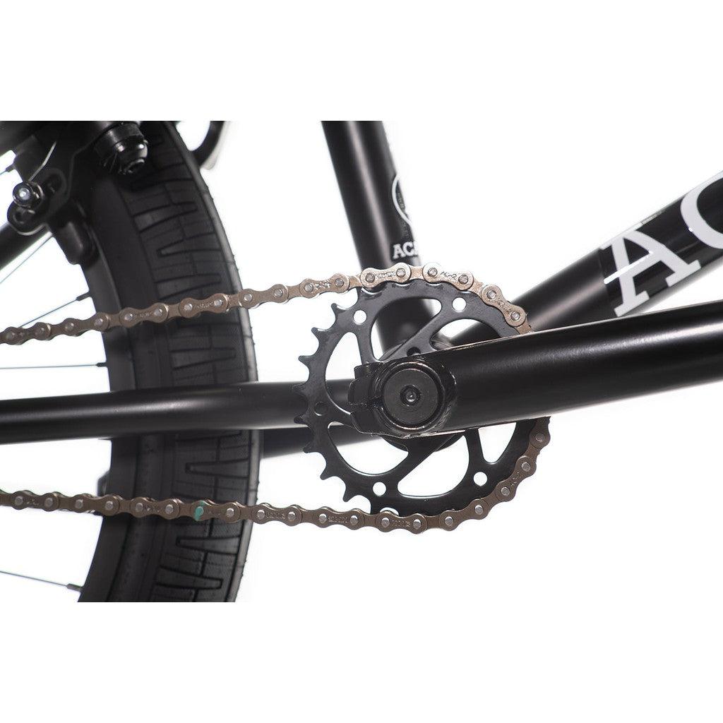 Close-up of the rear gear and chain of the Academy Trooper 20 Inch Bike attached to the wheel hub, featuring a Hi-Ten steel frame. The black frame and tire of the bike are partially visible.