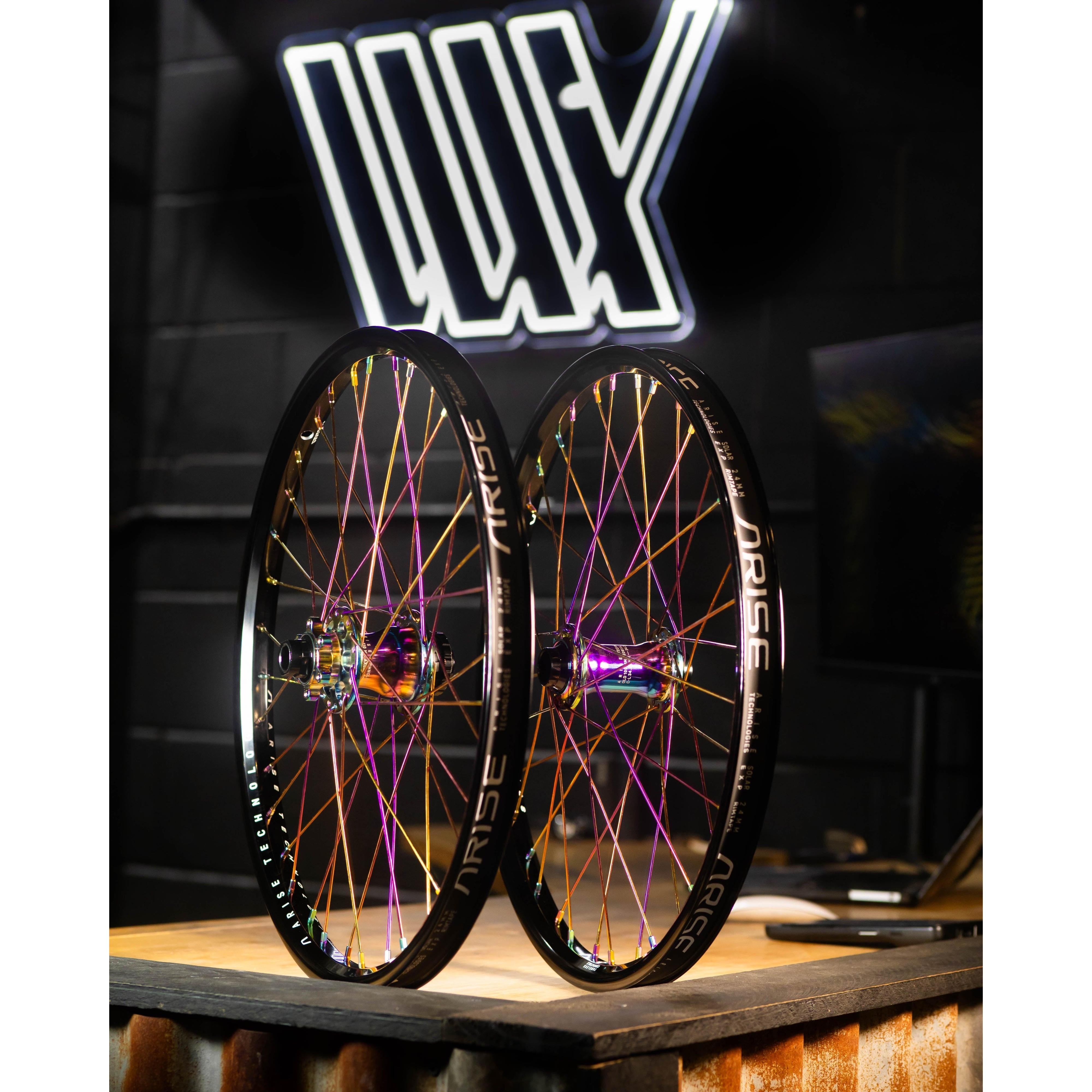 Two brightly lit bicycle wheels from the Arise Echo Oilslick Pro 20 Inch Wheelset stand upright on a wooden surface, their colorful spokes sparkling. A neon sign with stylized letters is visible in the background, suggesting high engagement for riders seeking style and performance.