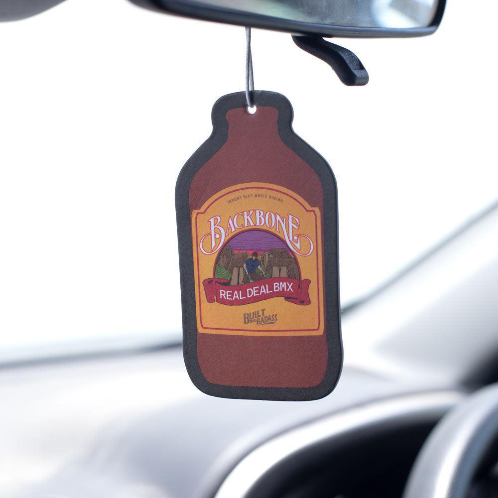 A Backbone Air Freshener with a spicy pineapple scent, shaped like a brown bottle and labeled "Backbone Real Deal BBQ," hangs from a car's rearview mirror.