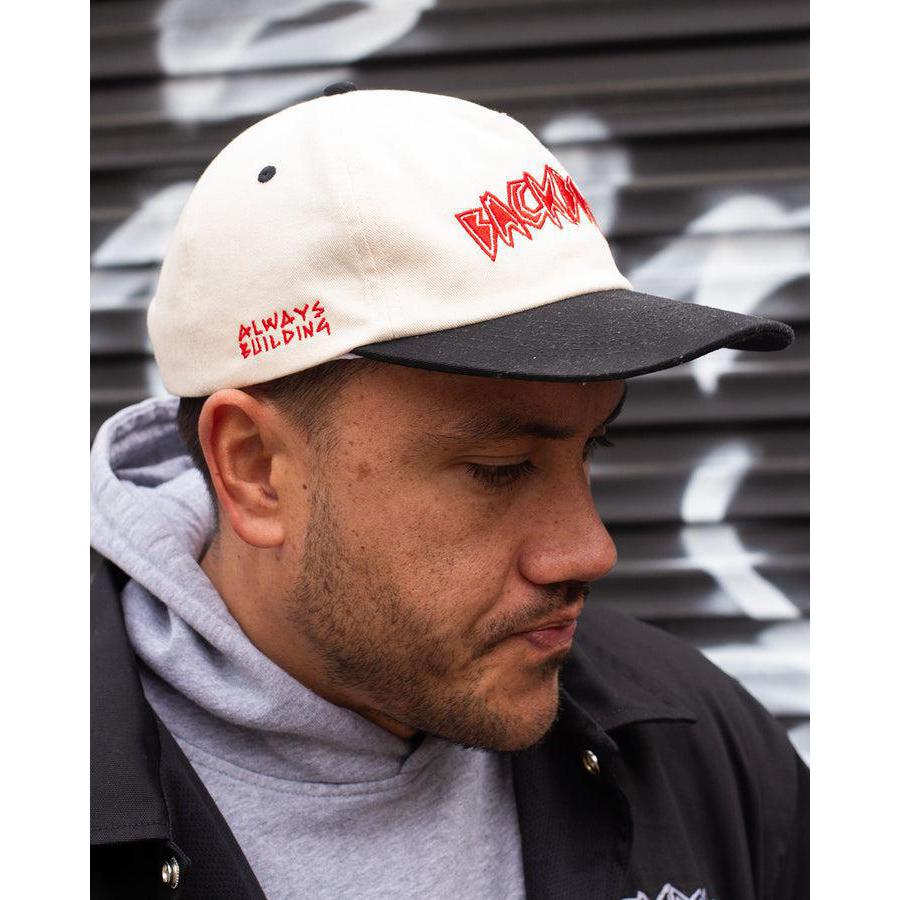 A man in a Backbone Always Building Hat, featuring a white and black design with an embroidered logo, pairs it with a gray hoodie and black jacket, posing effortlessly against a corrugated metal backdrop to capture the casual essence of Backbone BMX.