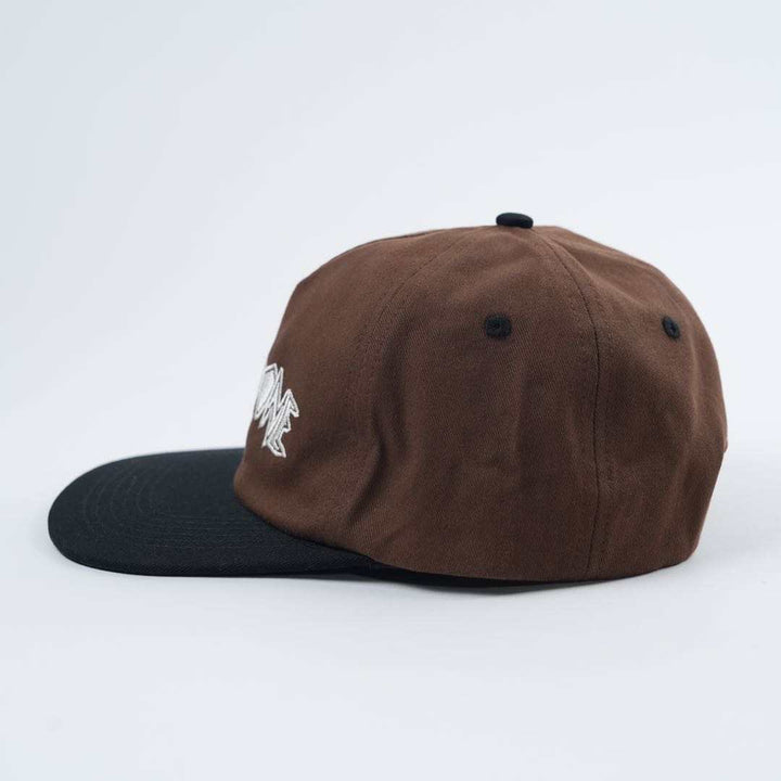 The Backbone Always Building Hat, a brown baseball cap with an embroidered logo and black brim, seen from the side on a plain light background, embodies the Backbone BMX spirit.