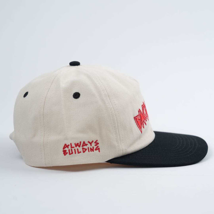 The Backbone Always Building Hat is an unstructured beige baseball cap with a black brim, featuring an embroidered logo and "ALWAYS BUILDING" in bold red text on the side, ideal for Backbone BMX fans.