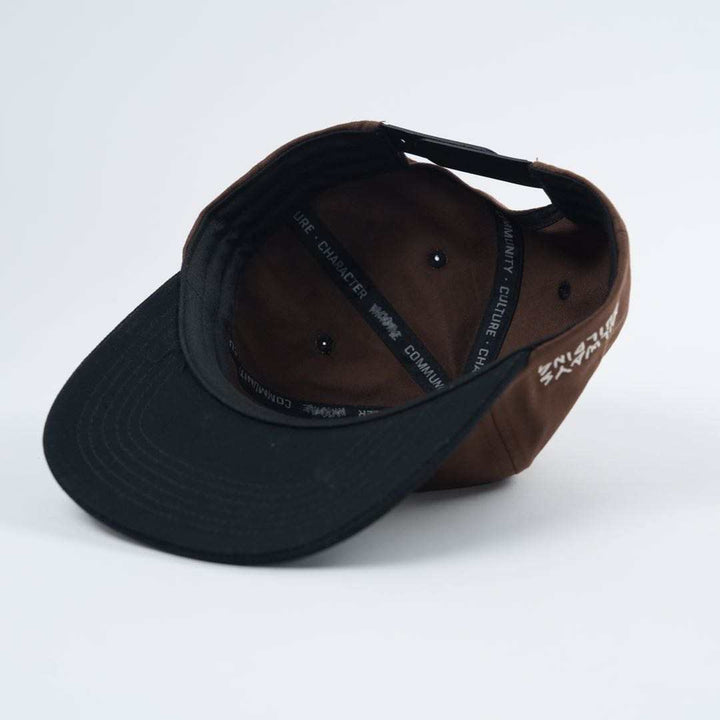 The Backbone Always Building Hat, a brown and black baseball cap, is flipped upside down on a white surface showing an embroidered logo along the brim's interior with subtle Backbone BMX flair.