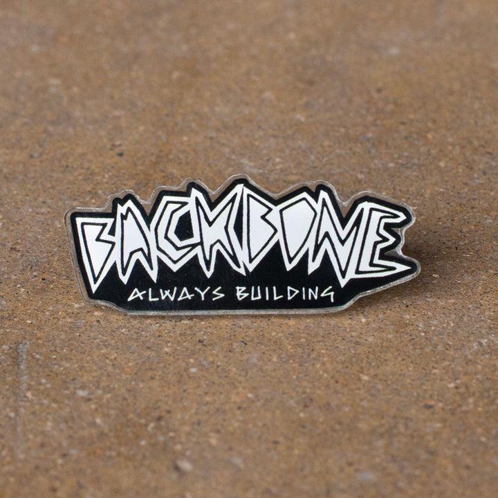 The Backbone Always Building Pin showcases graffiti-style "BACKBONE" and "ALWAYS BUILDING" text on a textured brown background. This pin symbolizes resilience, making it ideal for softgoods lovers and unique pin collectors, representing steadfast dedication.