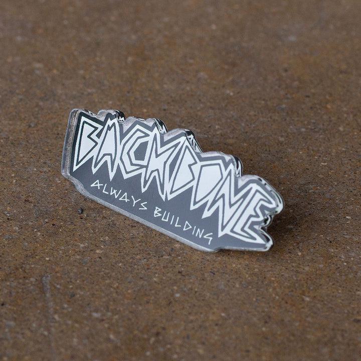 The Backbone Always Building Pin gleams on the textured surface, its bold, angular lettering showcasing Backbone's craftsmanship in pins and softgoods.