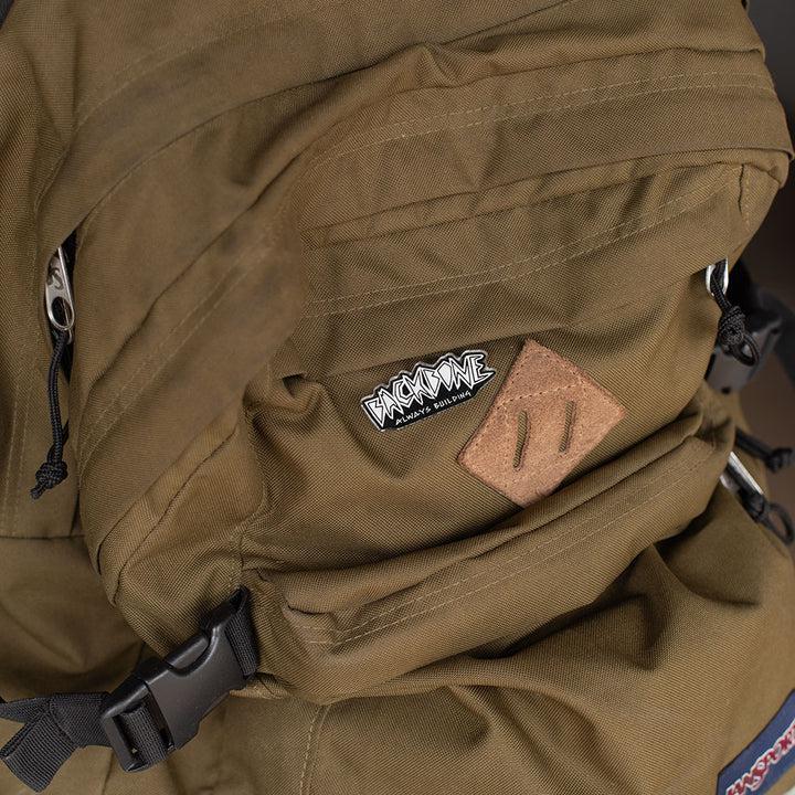 A close-up of a brown backpack highlights a black and white "Backbone Always Building Pin" logo patch, emphasizing its craftsmanship in softgoods.
