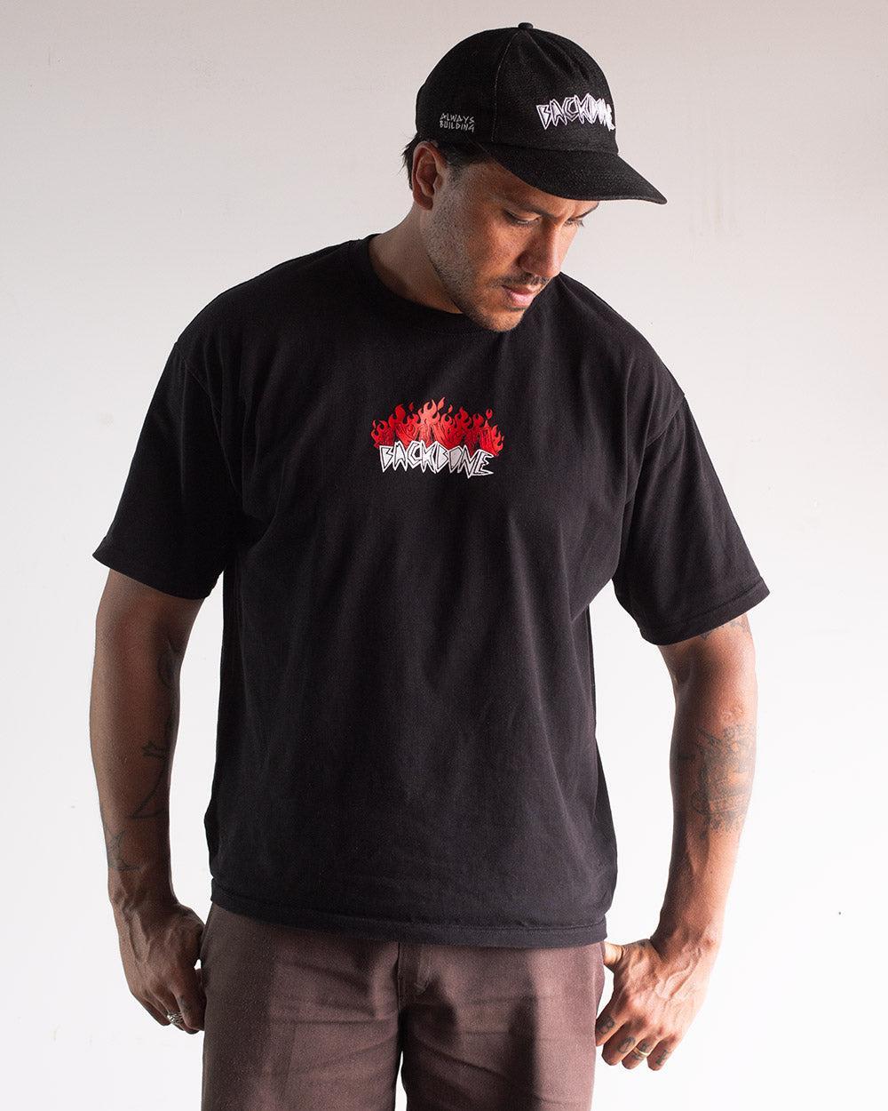 Man wearing the Backbone Fire Burning T-Shirt—a premium cotton, heavyweight tee featuring a red and white "BROOKLYN" graphic—paired with a matching black cap, standing against a plain background.