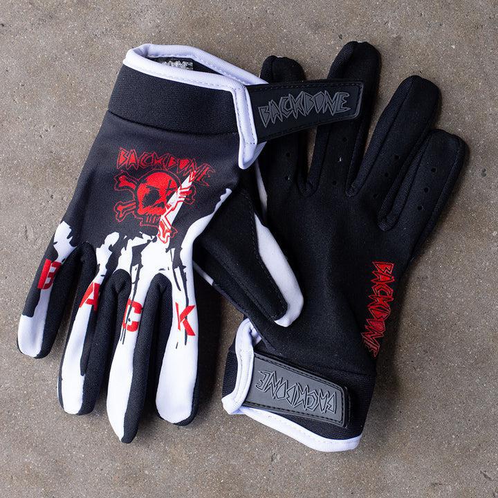 The Backbone Gnar Drip Adult Gloves are black and white with a red skull and crossbones logo, made from lightweight spandex, and strikingly placed on a textured surface.