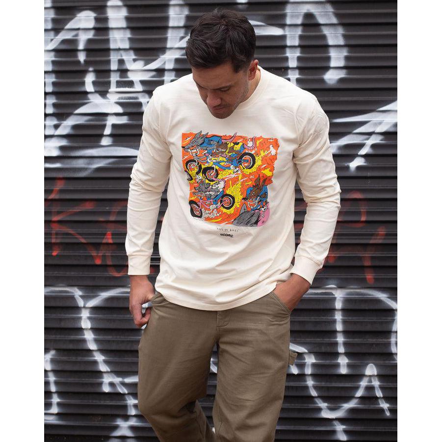 A man in a Backbone Rats On Bikes Long Sleeve Shirt by Zachary Jones-Peni and khaki pants stands against a graffiti-covered wall, looking down with one hand in his pocket. Made from 100% combed cotton, the shirt complements the urban backdrop perfectly.