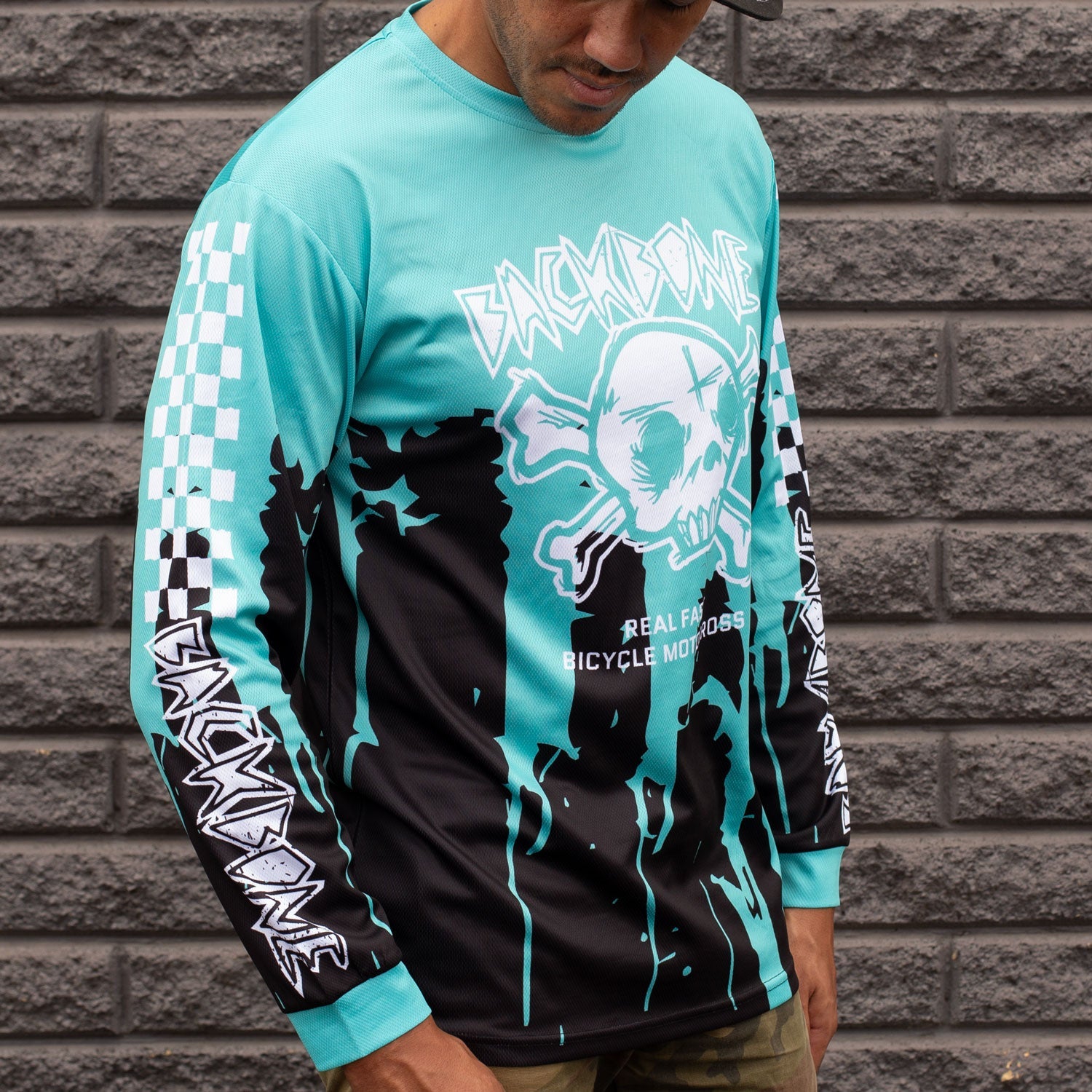 An individual stands against a brick wall wearing the Backbone Real Fast Race Jersey / Gnar Drip, showcasing turquoise and black long sleeves with a skull and crossbones design, customized sublimated detail, text, and checkered patterns.
