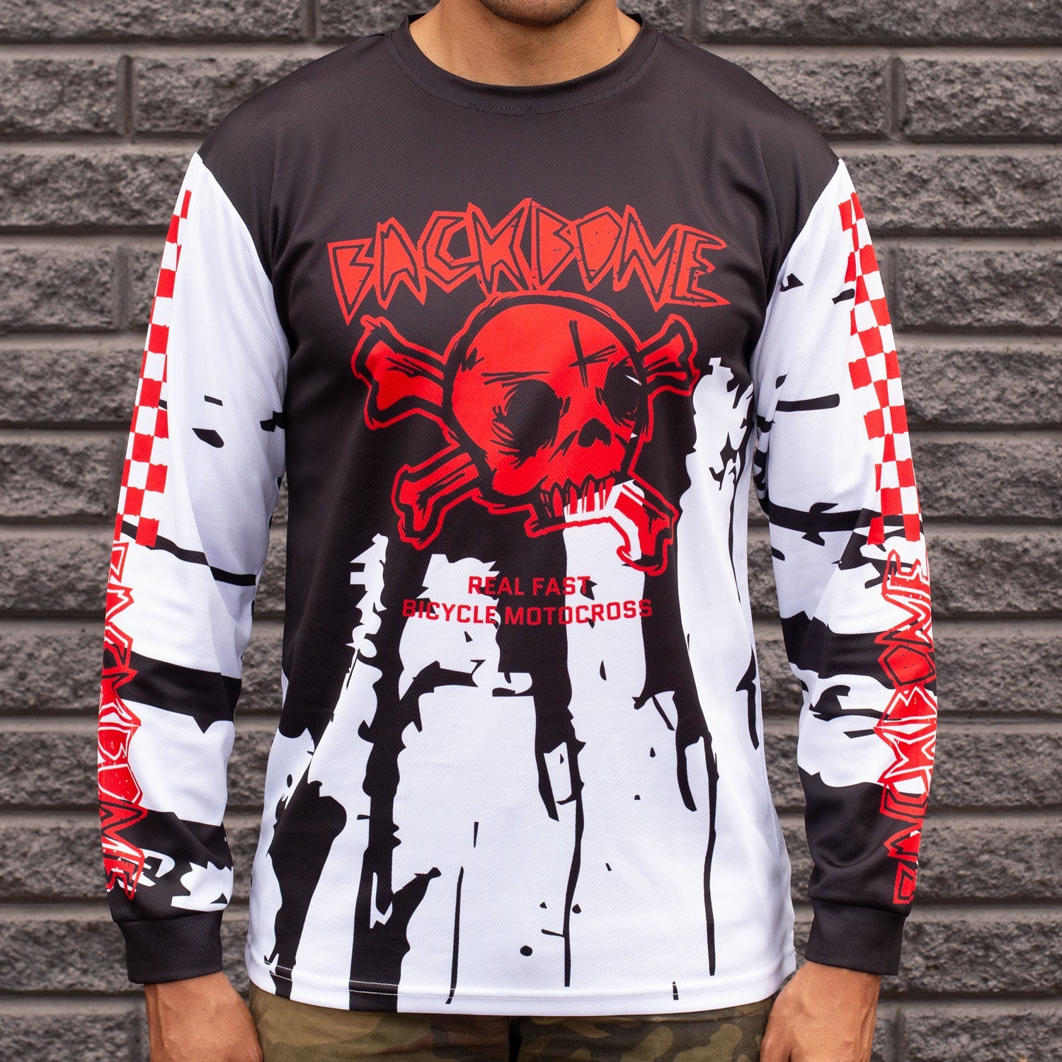 A person wears the Backbone Real Fast Race Jersey / Gnar Drip (Youth), showcasing a red skull, black and white splatter, and text "Backbone" and "Real Fast Bicycle Motocross," in custom sublimated polyester, standing against a gray brick wall.