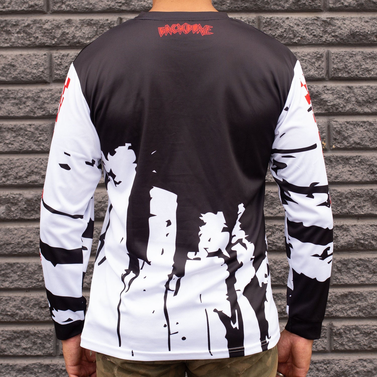Someone displays the Backbone Real Fast Race Jersey / Gnar Drip (Youth), a custom sublimated long-sleeve shirt with BACKBONE FIT's black and white design and bold red text on the upper back, against a gray brick wall backdrop.