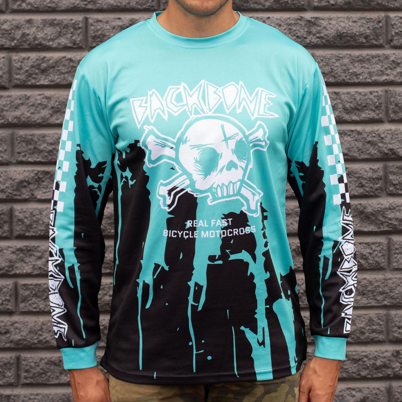 A person wearing the Backbone Real Fast Race Jersey in Gnar Drip, with a teal design featuring a skull graphic and "Real Fast Bicycle Motocross" text, stands against a gray brick wall.