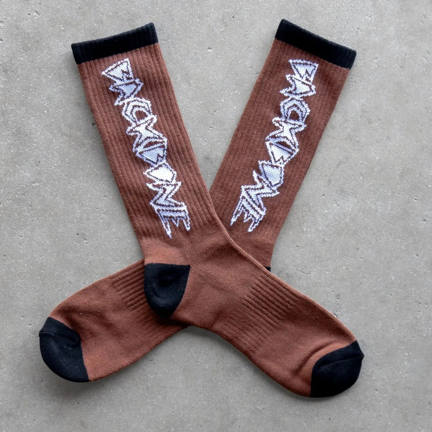 The Backbone Crew Socks are buttery soft, brown mid-shin socks with black toes, heels, and cuffs, featuring a white zigzag design along the sides and displayed on a grey surface.