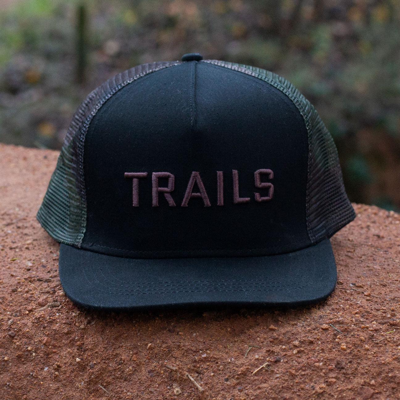 The Backbone Trails Trucker Hat is a black trucker hat with "TRAILS" embroidered in 3D on the front, shown outdoors on a brown surface.