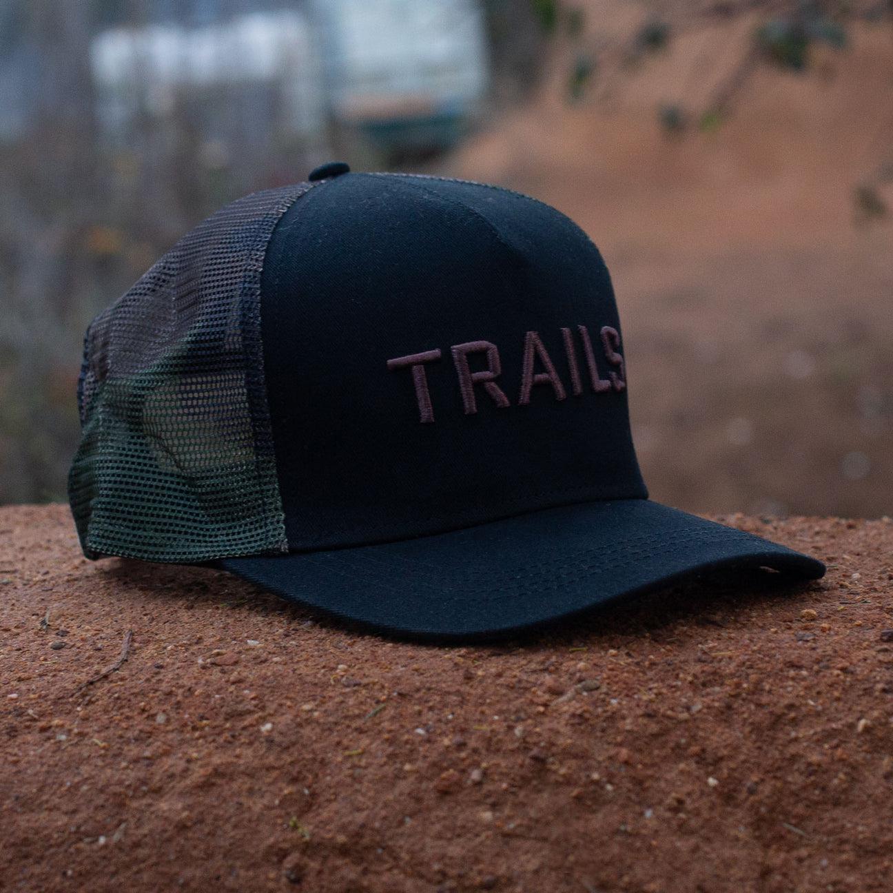 The Backbone Trails Trucker Hat, featuring "TRAILS" in 3D embroidery, sits on a brown surface outdoors, embodying the essence of bicycle motocross adventures.