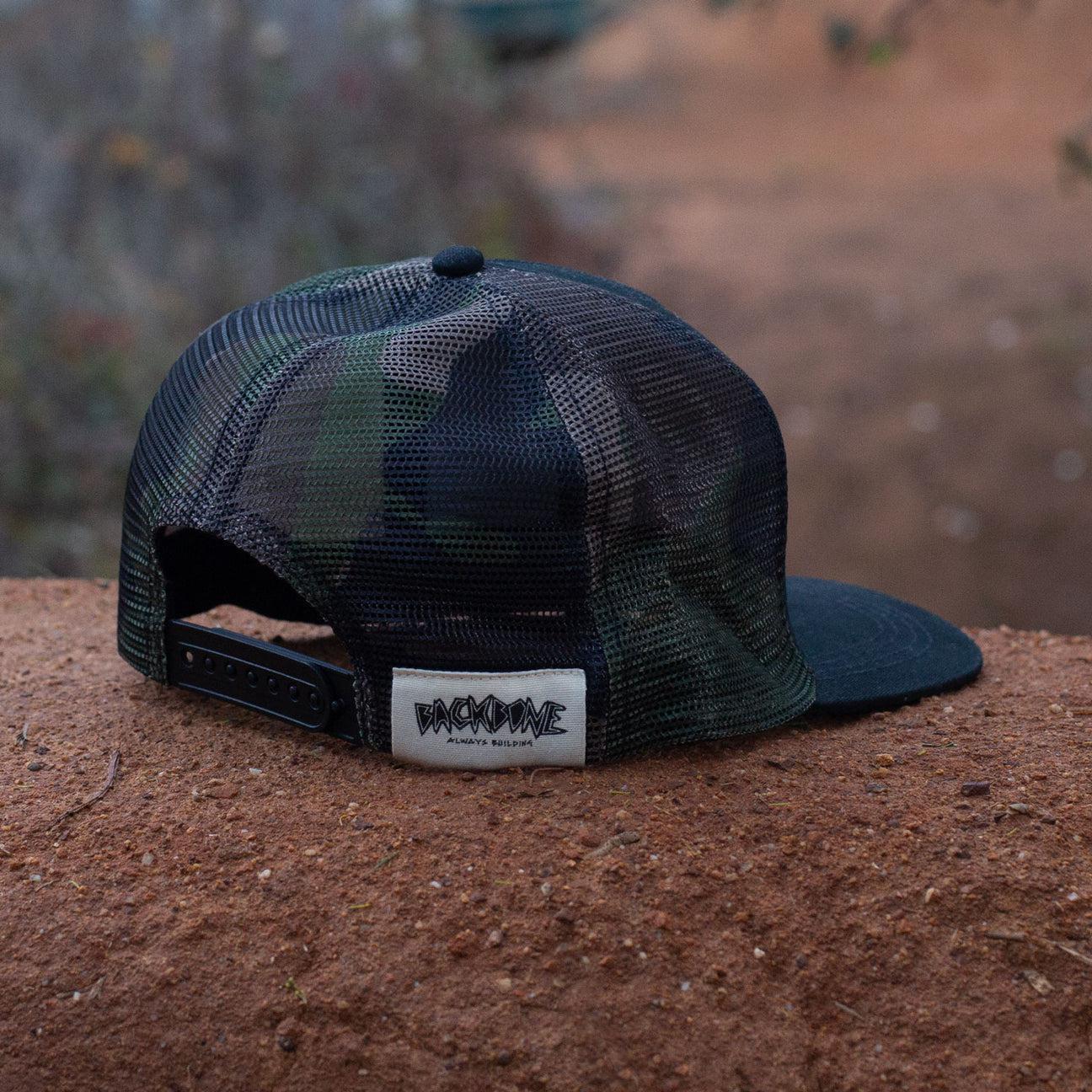 The Backbone Trails Trucker Hat, a camouflage snapback with mesh back, showcases 3D embroidery on a white patch featuring black graphic text, displayed on a textured outdoor surface.