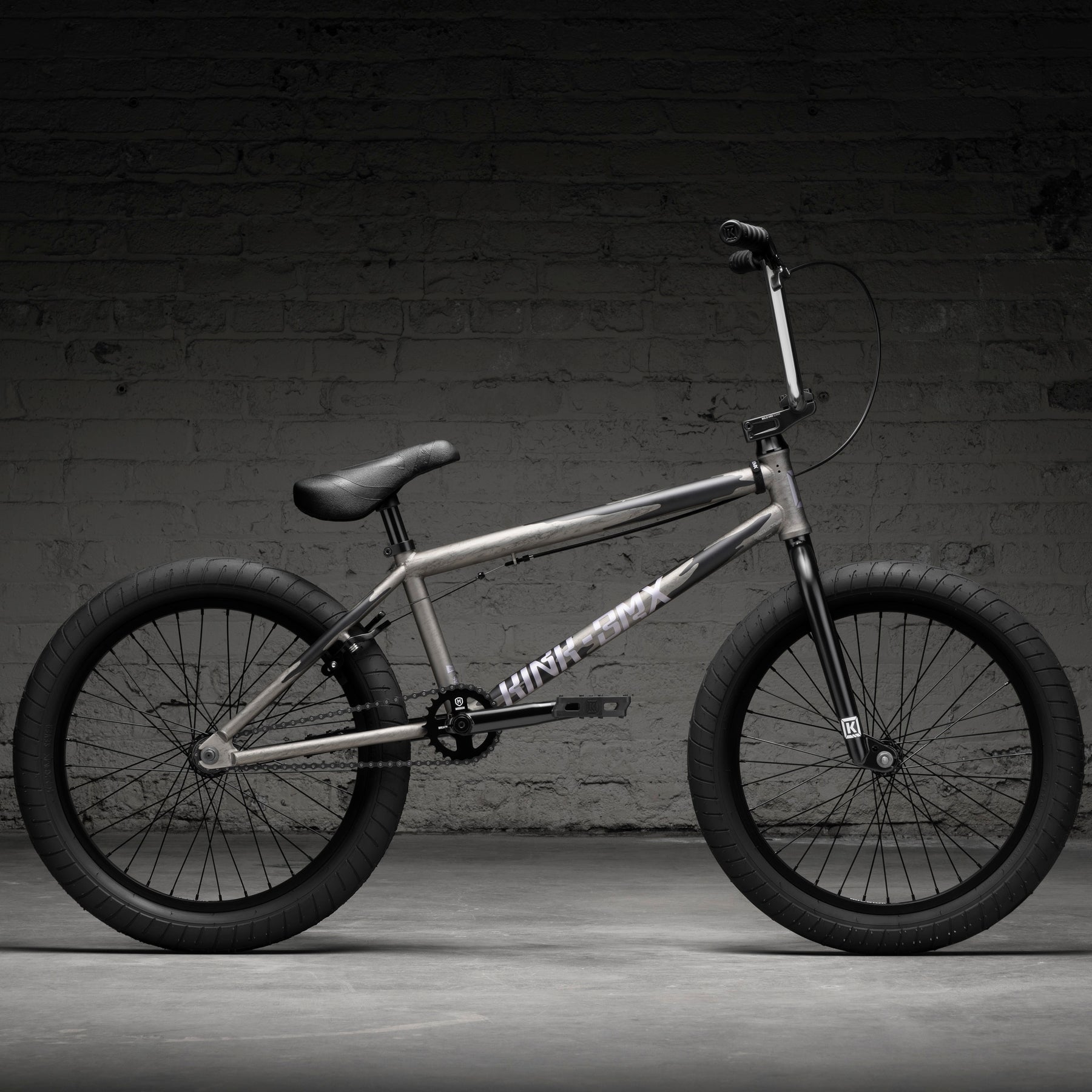 The Kink Launch LTD 20 Inch Bike is a limited edition BMX with a matte gray frame, black handlebars, seat, tires, and pedals, plus smooth sealed bearings. Shown against a dark brick wall background.