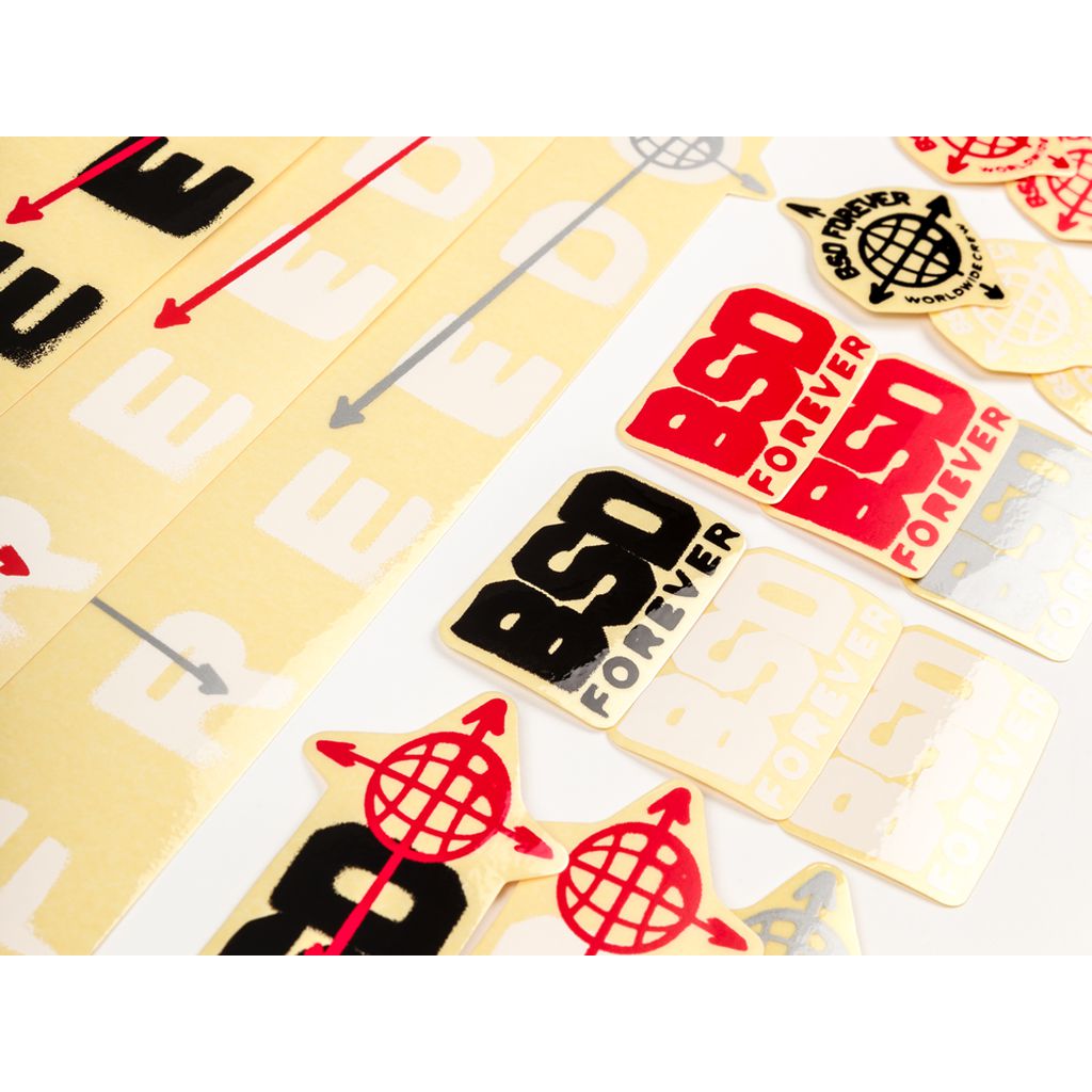 Stickers are arranged on a white surface. Some feature the text "BSD FOREVER" in red and black, while others depict red arrows and a globe. This fresh assortment is part of the new BSD Freedom Frame 2019 Decal Set.
