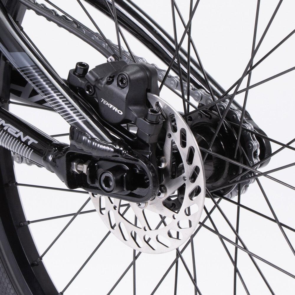 Close-up of the rear disc brake mechanism on the 2024 Chase Element Pro+ Cruiser Bike, featuring a Tektro brake caliper and a silver rotor with a perforated design. The Ikon carbon forks add an extra touch of performance to this sleek machine.