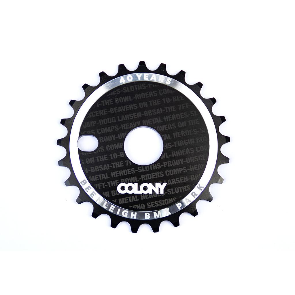 The Colony Beenleigh 40 Years CD Sprocket 25T, in sleek black and silver, commemorates the Colony Beenleigh skatepark's 40th anniversary with etched text and precise cutouts for mounting, celebrating a legacy of style and precision.