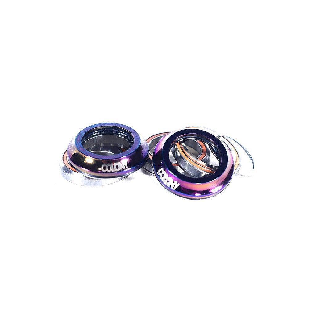 A set of colored bicycle headset parts, including two Cane Creek style cups labeled "Colony Integrated Headset," one in iridescent purple and blue, and one silver, featuring high quality cartridge bearings against a white background.