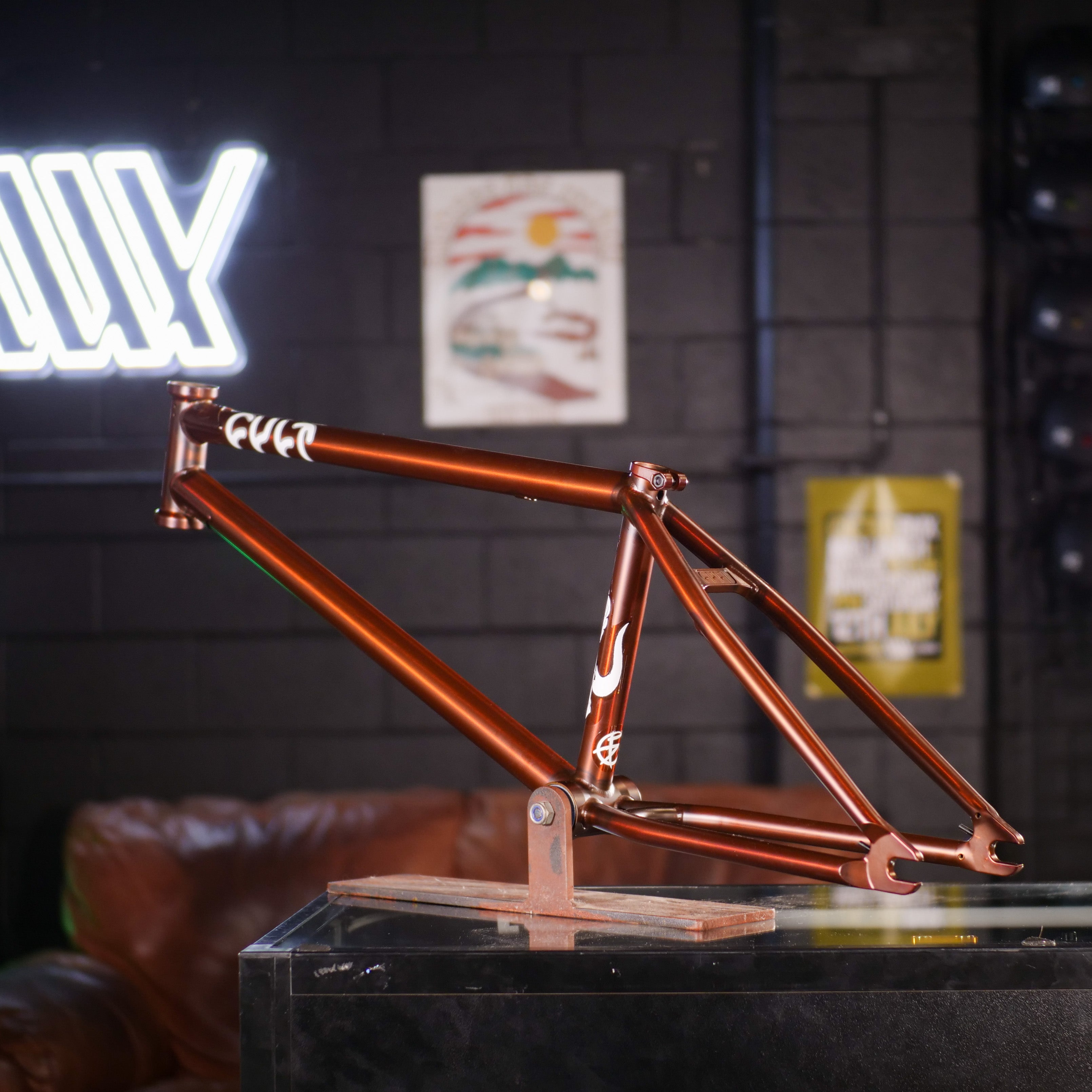 The Cult Dak Frame / Pat Freyne Signature—a metallic bronze BMX frame with 100% cult classic tubing—is showcased indoors, standing out against a backdrop of posters and a neon sign on a dark wall.