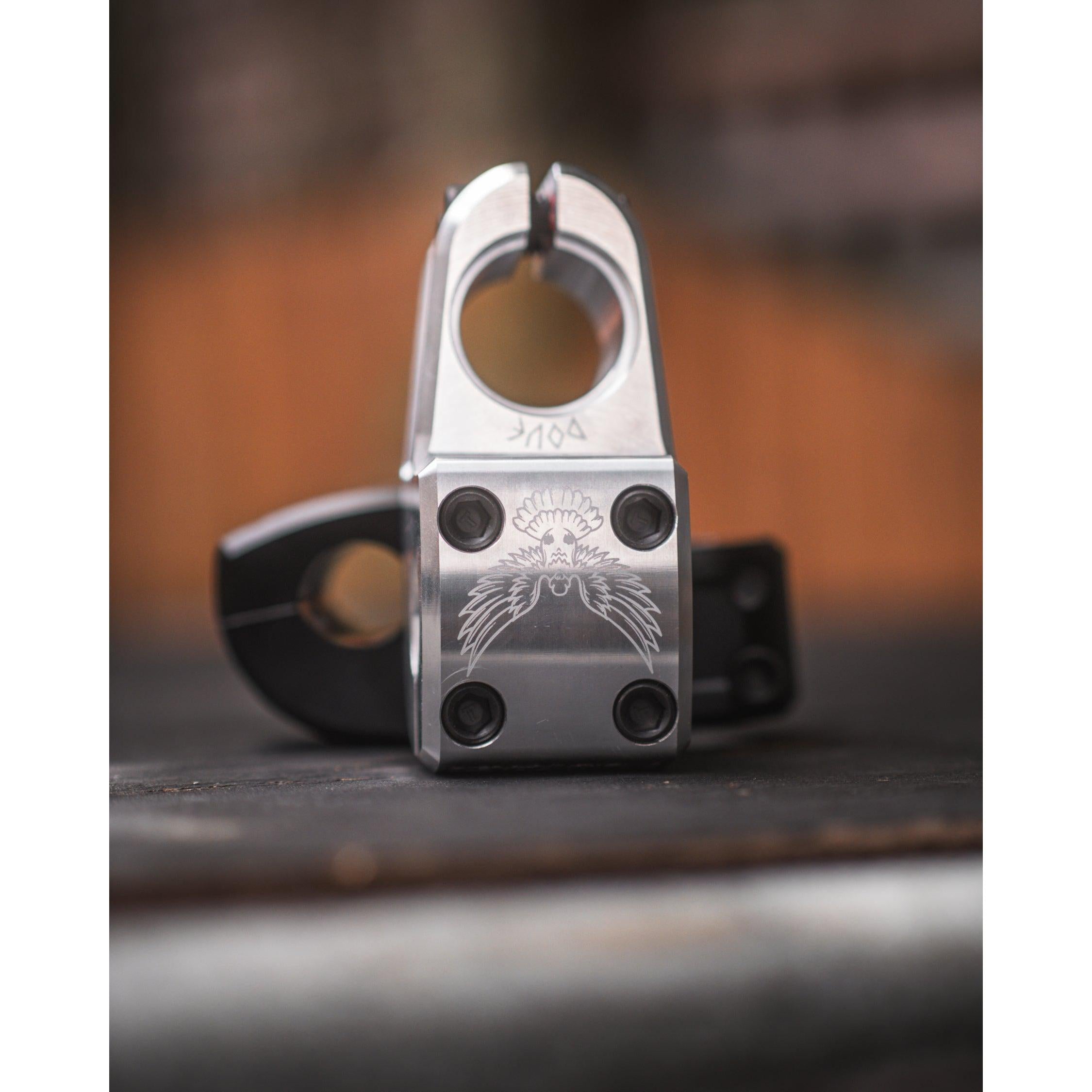 Close-up of the Demolition Josh Dove Stem with a stylized eagle engraving. Four screws ensure durability, while the blurred background in warm tones highlights its intricate craftsmanship.